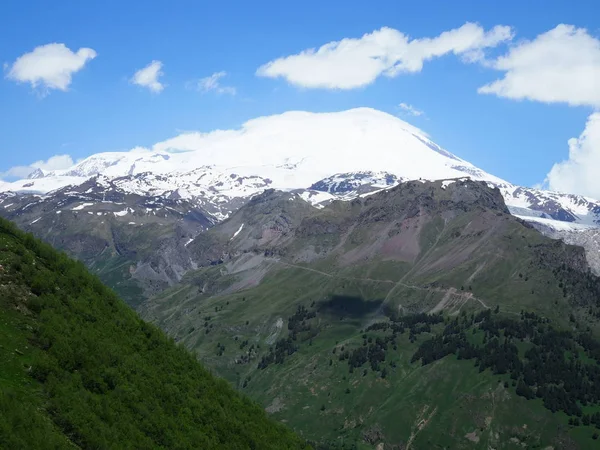 Great nature mountain range. Amazing perspective of caucasian snow mountain or volcano Elbrus with green fields, blue sky background. Elbrus landscape view - the highest peak of Russia and Europe