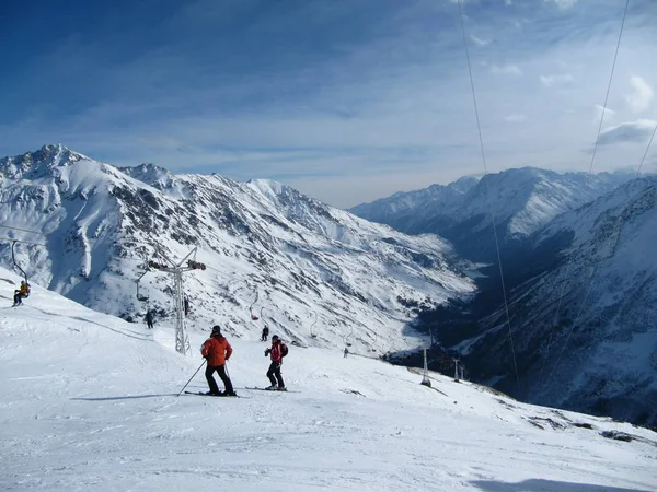 Лыжник стоит на склоне. Девушка в светлом костюме, шлем и маска в лыжах, чтобы кататься на лыжах. На заднем плане заснеженные горы, лыжники и подъемники. Кавказские горы, Эльбрус, Россия - февраль 2010 года — стоковое фото