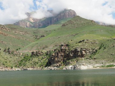 Kayalık dağların tepelerini beyaz bulutlar kaplıyor. berrak dağlı gölün yanında berrak su ve dağlarla Alp çayırları.
