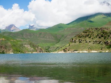 Kayalık dağların tepelerini beyaz bulutlar kaplıyor. berrak dağlı gölün yanında berrak su ve dağlarla Alp çayırları.