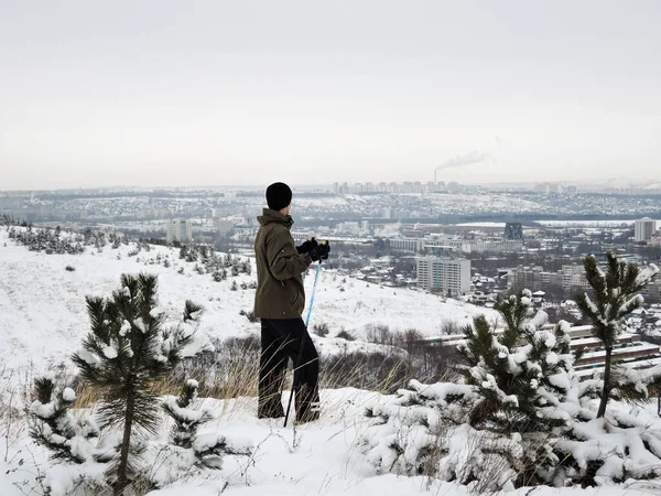 Mannelijke skiër staat aan de rand van het bos en kijkt naar het stedelijke landschap van de stad en de pijpen met rook die het milieu vervuilen. concept van downshifting. Rusland, Saratov - januari 2020 — Stockfoto