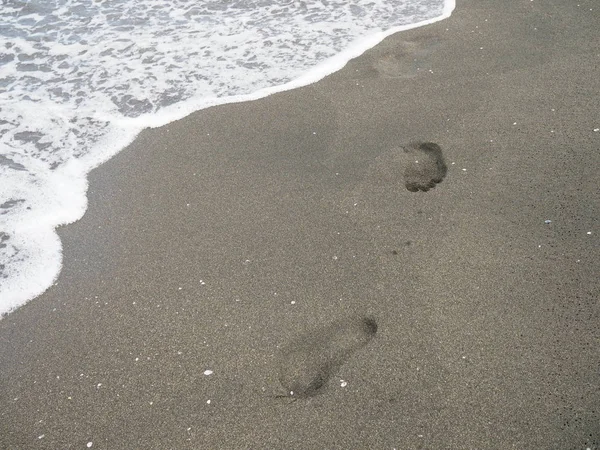Fußabdrücke an einem schwarzen Sandstrand mit Tilt-Shift-Unschärfe. Surfen im Hintergrund. Konzept für Einsamkeit, Einsamkeit, Depression. — Stockfoto