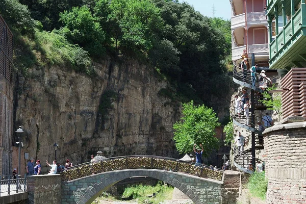 Georgia, Tbilisi - June 2019. Pastel-colored homes sit on a cliff above a small stream accessed by a spiral staircase and crossed by an arched stone bridge on a sunny day. — 图库照片