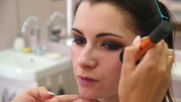 Maquillaje profesional artista con un cepillo aplica rubor en la cara de una mujer de ojos marrones. Maquillaje artista haciendo maquillaje para la joven, primer plano . — Vídeo de stock