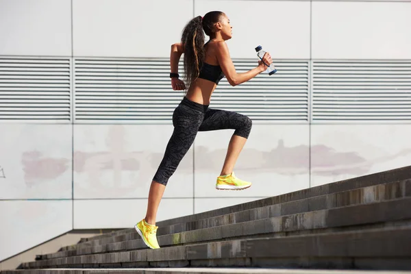 Mujer atlética corriendo hasta las escaleras de piedra con botella de agua en la ciudad con rastreador de fitness — Foto de Stock