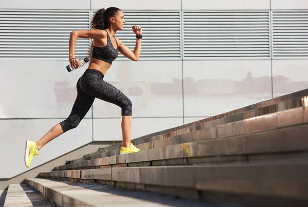 Mujer atlética corriendo hasta las escaleras de piedra con botella de agua en la ciudad con rastreador de fitness — Foto de Stock