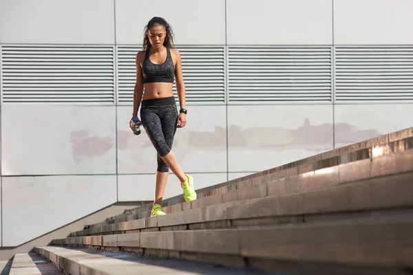 Tiro ao ar livre de uma jovem mulher de pé em uma escada com garrafa de água e olhando para longe. Fitness fêmea antes de uma corrida . — Fotografia de Stock