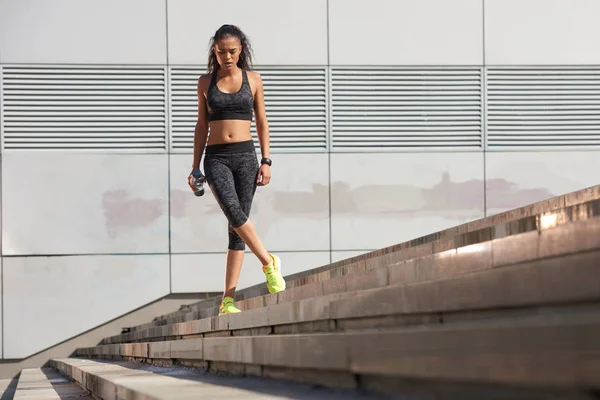 Foto al aire libre de una mujer joven parada en una escalera con una botella de agua y mirando hacia otro lado. Fitness femenina antes de una carrera . — Foto de Stock