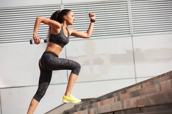 Mujer atlética corriendo hasta las escaleras de piedra con botella de agua en la ciudad con rastreador de fitness — Foto de Stock