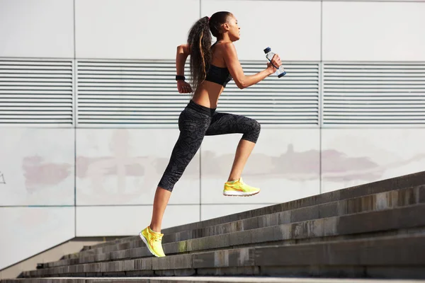 Atletisk kvinna kör till sten trappan med flaska vatten i City med fitness tracker Royaltyfria Stockfoton