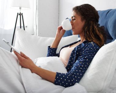 Bir tablet bilgisayar kullanma ve içme kahve sırasında yatakta genç casual kadın