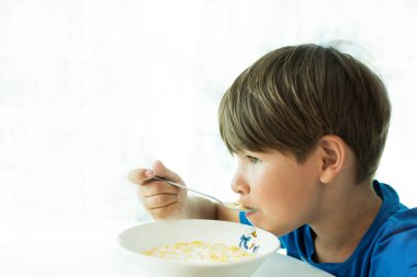 Mavi tişörtlü bir çocuk kahvaltısını beyaz tabakta yulaf ezmesi ve sütle yapar.