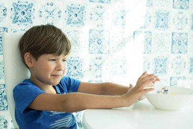 a boy in a blue T-shirt has breakfast with oatmeal and milk in a white plate, space for text, poor appetite, the child does not eat clipart