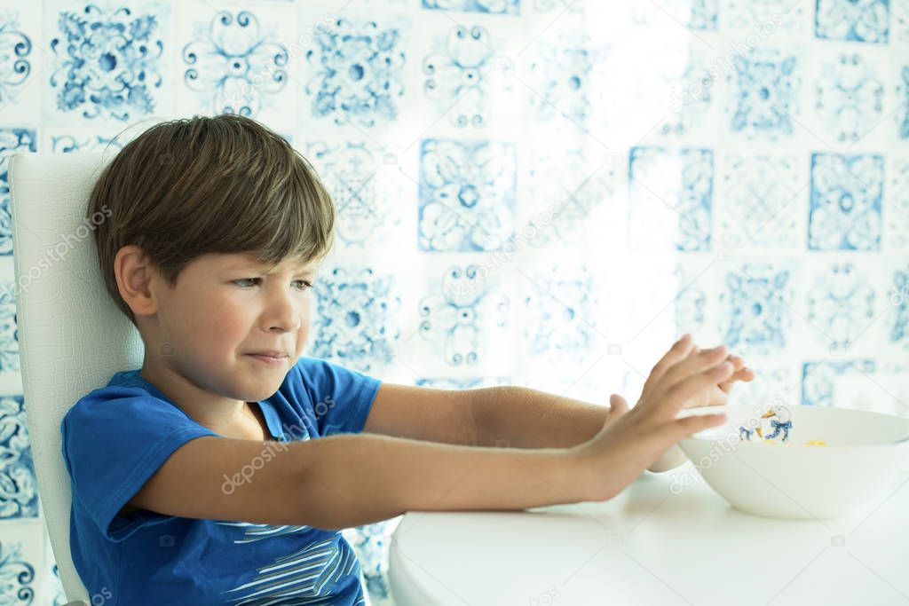 a boy in a blue T-shirt has breakfast with oatmeal and milk in a white plate, space for text, poor appetite, the child does not eat