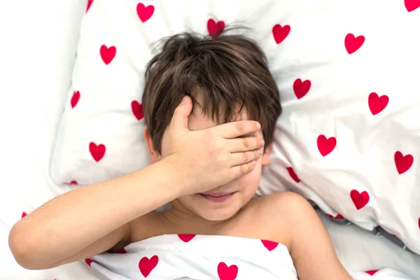 O menino jaz em uma cama com corações vermelhos, seu rosto em suas mãos. emoções sem rosto. cor branca, vista superior — Fotografia de Stock