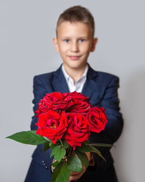 A boy in a blue suit holds a bouquet of red roses. Grey background. 