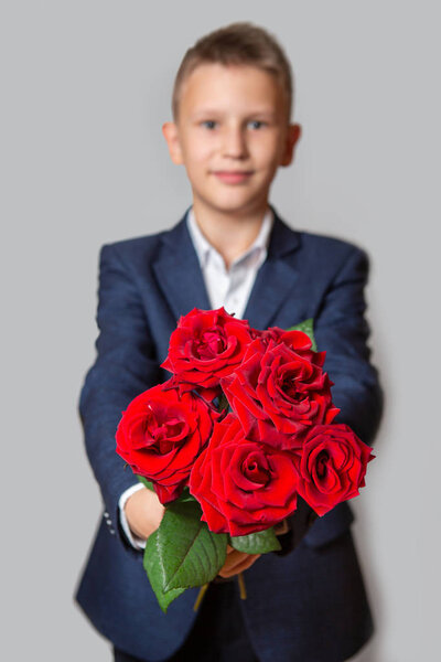 A boy of Caucasian appearance in a blue suit holds a bouquet of red roses. Grey background. 