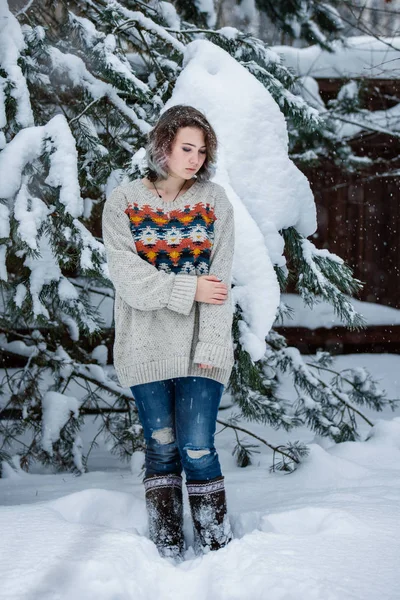 Uma menina de aparência caucasiana em uma camisola e jeans em um fundo de árvores nevadas. Dia de inverno nevado. Um passeio divertido . — Fotografia de Stock