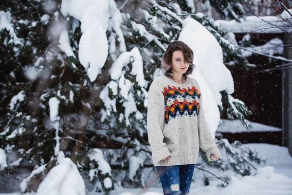 Uma menina sorridente de aparência caucasiana em uma camisola e jeans em um fundo de árvores nevadas. Dia de inverno nevado. Um passeio divertido . — Fotografia de Stock