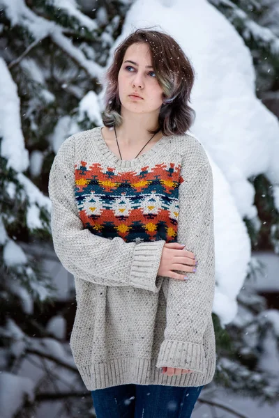 A girl of Caucasian appearance in a sweater and jeans on a background of snowy trees. Snowy winter day. Pensive girl.