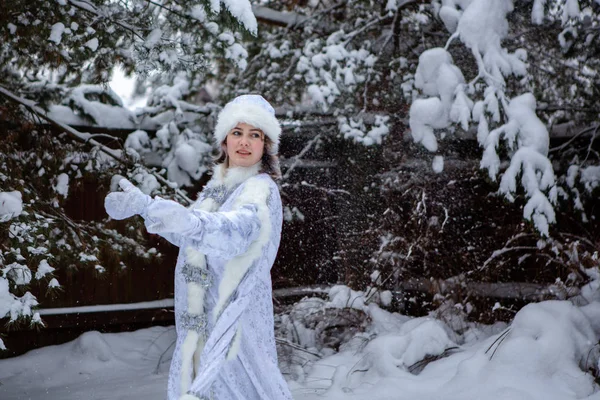 Uma menina vestida como Donzela da Neve joga bolas de neve em um fundo de árvores nevadas. Natal e Ano Novo, inverno . — Fotografia de Stock