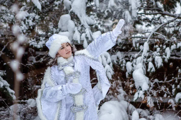 Ένα κορίτσι ντυμένο Χιονάτη ρίχνει χιονόμπαλες σε ένα φόντο χιονισμένων δέντρων. Χριστούγεννα και Πρωτοχρονιά, χειμώνας. — Φωτογραφία Αρχείου