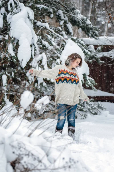 Uma menina sorridente de aparência caucasiana em uma camisola e jeans em um fundo de árvores nevadas. Dia de inverno nevado, nevasca . — Fotografia de Stock