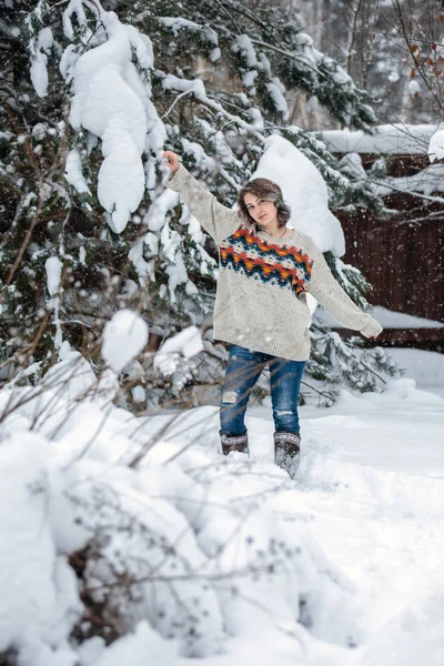 Uma menina sorridente de aparência caucasiana em uma camisola e jeans em um fundo de árvores nevadas. Dia de inverno nevado, nevasca . — Fotografia de Stock