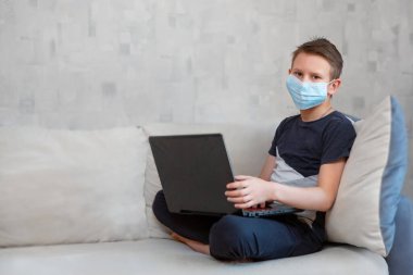 Tek kullanımlık maskeli dizüstü bilgisayarı olan bir çocuk koltukta ödev yapıyor. Karantinada uzaktan öğrenme. Ders kitapları ve defterler çocuğun yanındadır..