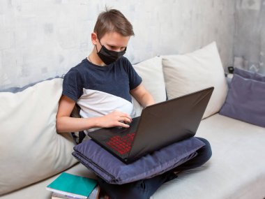 Tek kullanımlık maskeli dizüstü bilgisayarı olan bir çocuk koltukta ödev yapıyor. Karantinada uzaktan öğrenme. Ders kitapları ve defterler çocuğun yanındadır..