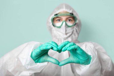 Güvenlik gözlüklü, maskeli ve takım elbiseli bir sağlık görevlisi kalp işareti yapar. Yeşil homojen arka plan. Uzayı kopyala Koronavirüsle yüzleşmek.