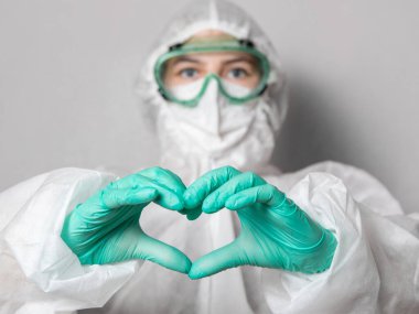 Güvenlik gözlüklü, maskeli ve takım elbiseli bir sağlık görevlisi kalp işareti yapar. Gri homojen arka plan. Uzayı kopyala Koronavirüsle yüzleşmek.
