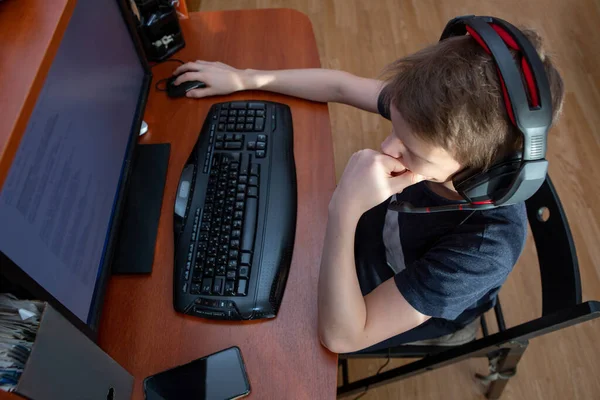 A boy in headphones in front of a computer does his homework. Distance learning in quarantine. Textbooks, notebooks  are on the table.