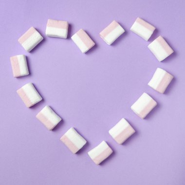 Beyaz ve pembe şekerlemeler. Mor bir arka planda marşmelovdan yapılmış kalp şekli. Tatlı günü. Düz yatıyordu.