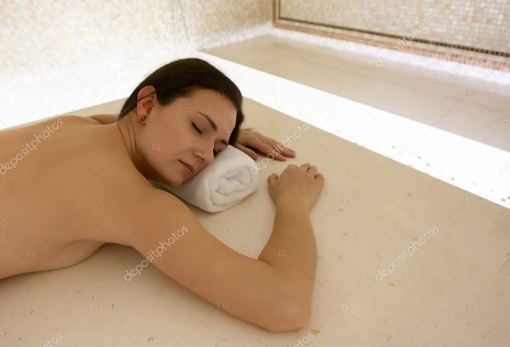 A young woman is relaxing in a traditional hammam, waiting for a Moroccan spa treatment and massage, lying on a marble bench with her eyes closed.