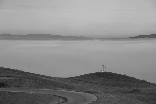 Black and white picture of small wooden cross on a hill overlooking a valley that is inundated by thick mist