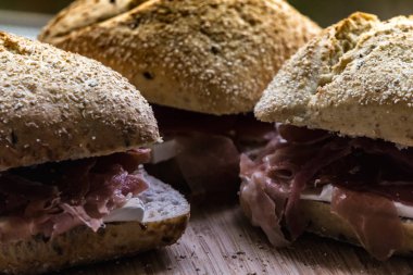 Tahta kesme tahtasının üzerinde kepekli ekmek, serrano jambonu ve peynirli üç sandviç ve küçük ekmek kırıntıları.