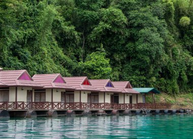 Cheow Lan Gölü 'nde yüzen güzel bungalovlar. Arkasında yeşil bir orman duvarı var.