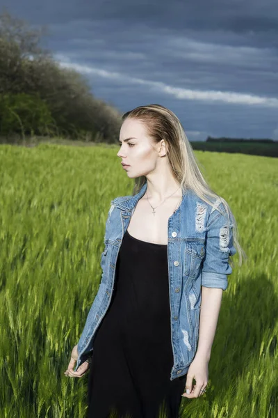 Menina loira bonita está de pé em um campo contra um fundo de nuvens . — Fotografia de Stock
