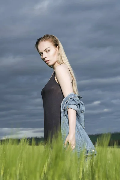 Menina loira bonita está de pé em um campo contra um fundo de nuvens . — Fotografia de Stock