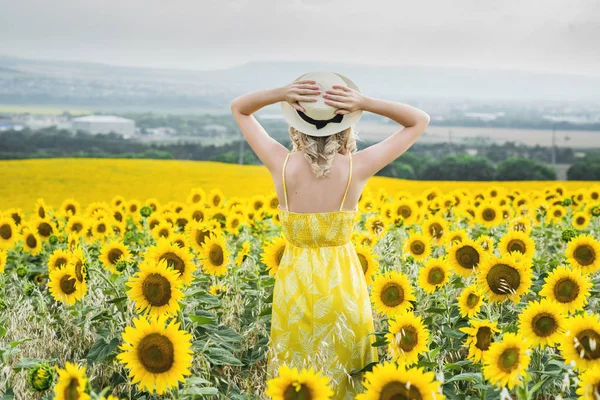 Молодая девушка стоит в поле подсолнухов, держа соломенную шляпу руками, вид сзади . — стоковое фото
