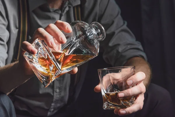 Close-up das mãos de um jovem que derrama álcool de um decantador em um copo, em um fundo escuro isolado — Fotografia de Stock