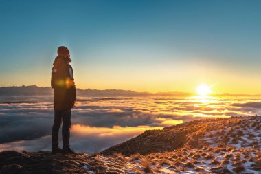 Bir adam yüksek dağlarda, gün batımında, bulutların üzerinde duruyor..