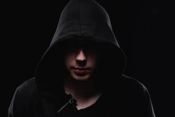 A man in a black hood on a black background, studio photography. The idea of mysticism, mystery, crime and deception