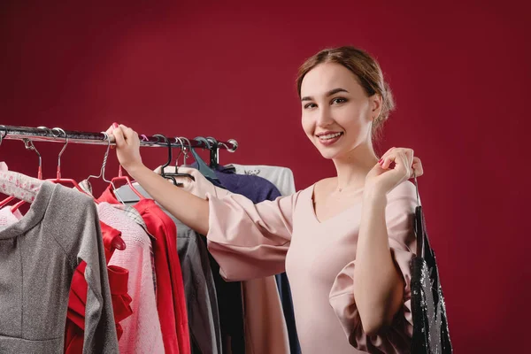 Menina bonita escolhe e experimenta roupas em um fundo vermelho. O conceito de escolher e comprar roupas, shopaholism e consumismo — Fotografia de Stock