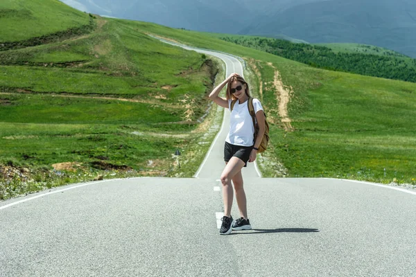 Une jeune fille avec un sac à dos se tient sur une route dans les montagnes. Le concept de l'auto-stop de voyage et de loisirs — Photo