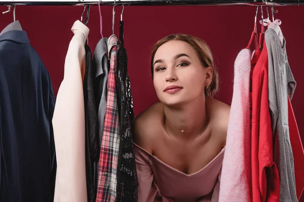 Menina bonita escolhe e experimenta roupas em um fundo vermelho. O conceito de escolher e comprar roupas, shopaholism e consumismo — Fotografia de Stock