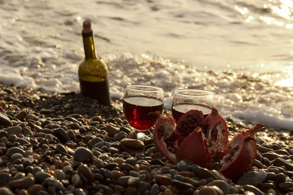 Ripe pomegranate fruit with a glass of wine and a bottle on the beach. Romantic day at the seaside.