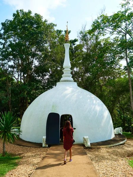 A female is walking towards a dome shaped building. The building is white in color. The building is located within the area of black house, an art museum in Chiang Rai Thailand.