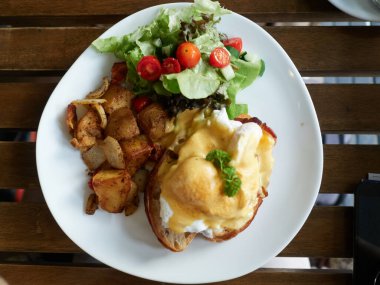 Büyük beyaz bir tabakta servis edilen Benedict usulü bir kahvaltı. İçinde biraz vişneli domates, fırında patates ve yapraklı sebze var..